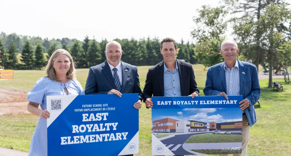 Four people stand in front of the new school site holding signs that depict the future buildings design.