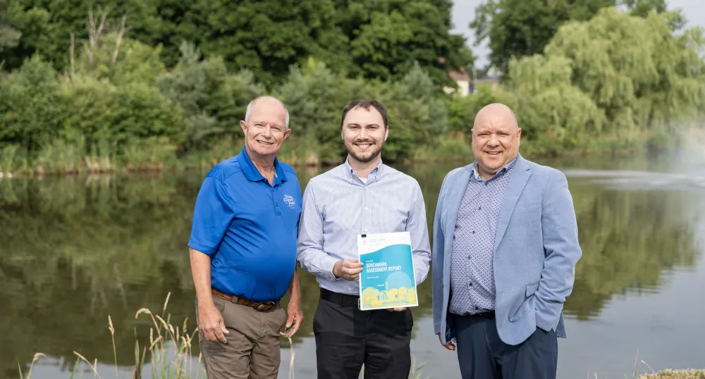 Three white men stand next to each other in front of a pond with a background of green trees.
