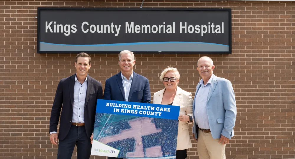 Three men and one woman stand in front of the Kings County Memorial Hospital sign while holding a poster of the newly acquired land.
