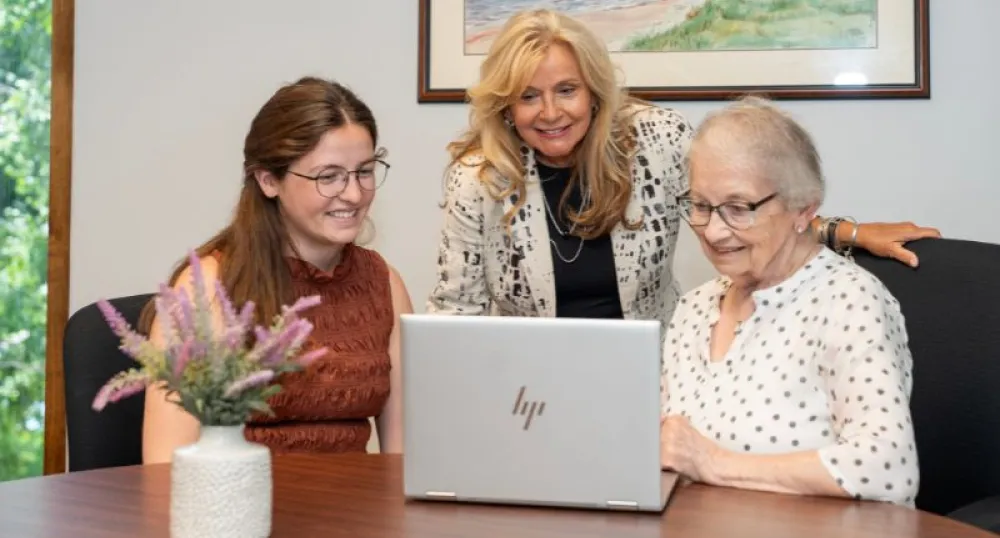 An elder woman and two other women gather around a computer screen.