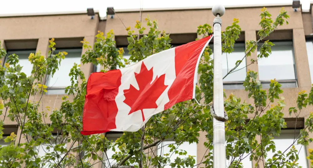 A Canadian flag blows in the wind with trees in the background.
