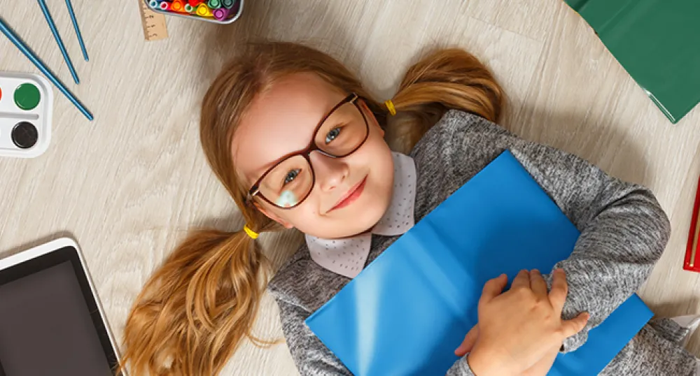 Young blonde girl with glasses laying on her back on the floor holding a book surrounded by books, pencils, paint and paint brushes
