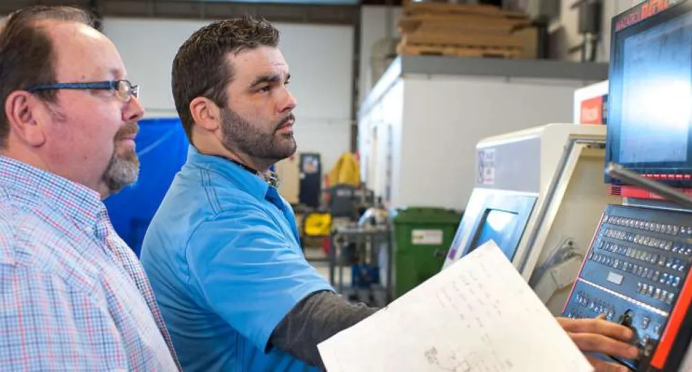 Tool and die maker looks on as a machinist sets up a CNC machine