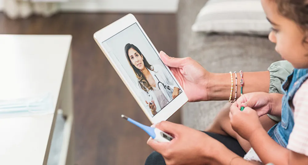 Child sitting on a mothers lap holding a thermometer and tablet speaking with a virtual care physician