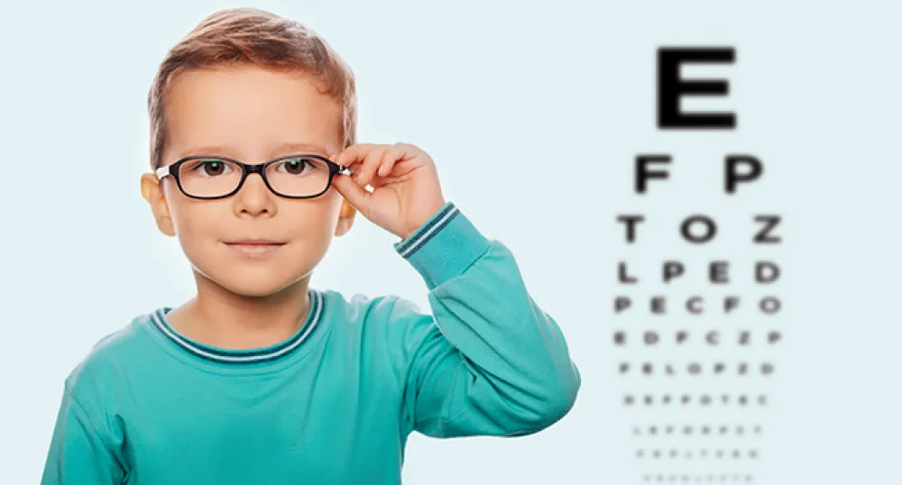 Young boy with glasses on, holds them with one hand to check his vision while an eye chart is the in the background