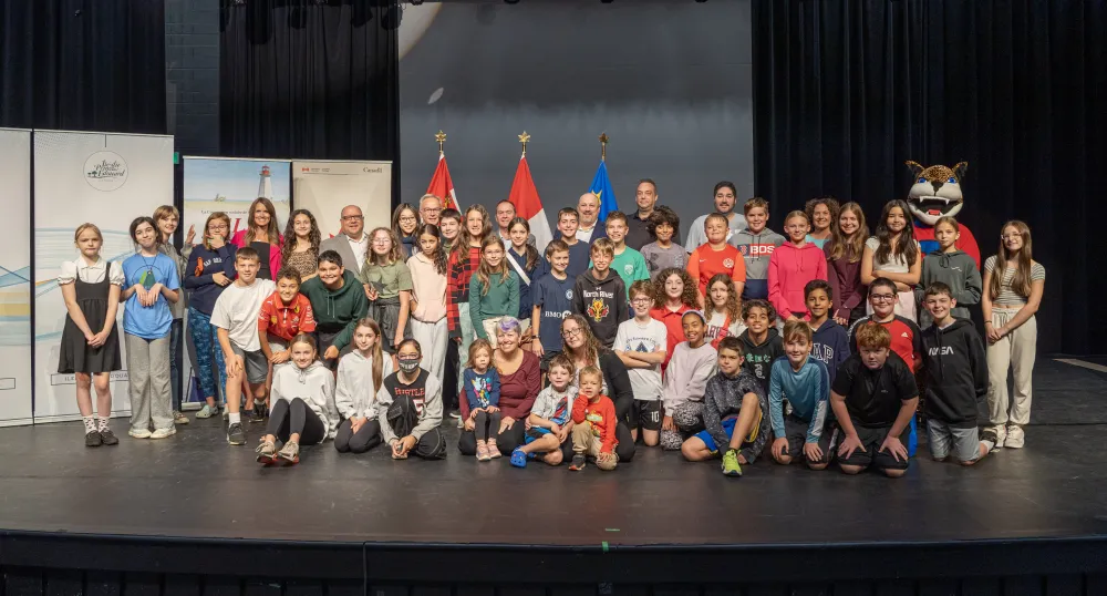 Important financement pour l’École François-Buote et le Carrefour de l’Isle-Saint-Jean roupe de jeunes étudiants et de quelques adultes sur une scène de spectacle avec des drapeaux en arrière-plan
