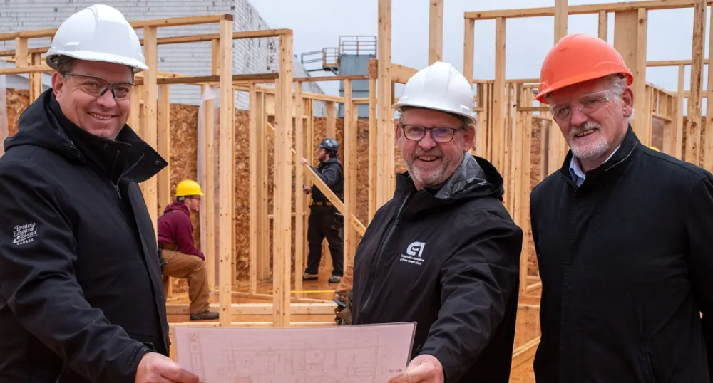 Province partners with Holland College and Construction Association to build tiny homes image of three people standing shoulder to shoulder holding design plans in front of a small b house under construction