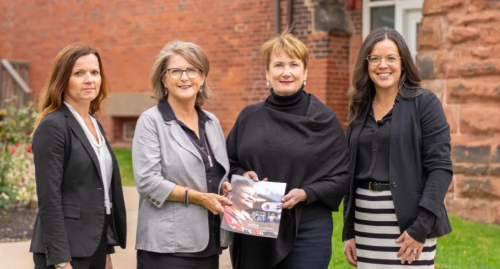 Long term care review identifies recommendations for enhancement four people standing shoulder to shoulder in front of a brick building with two of them holding a booklet