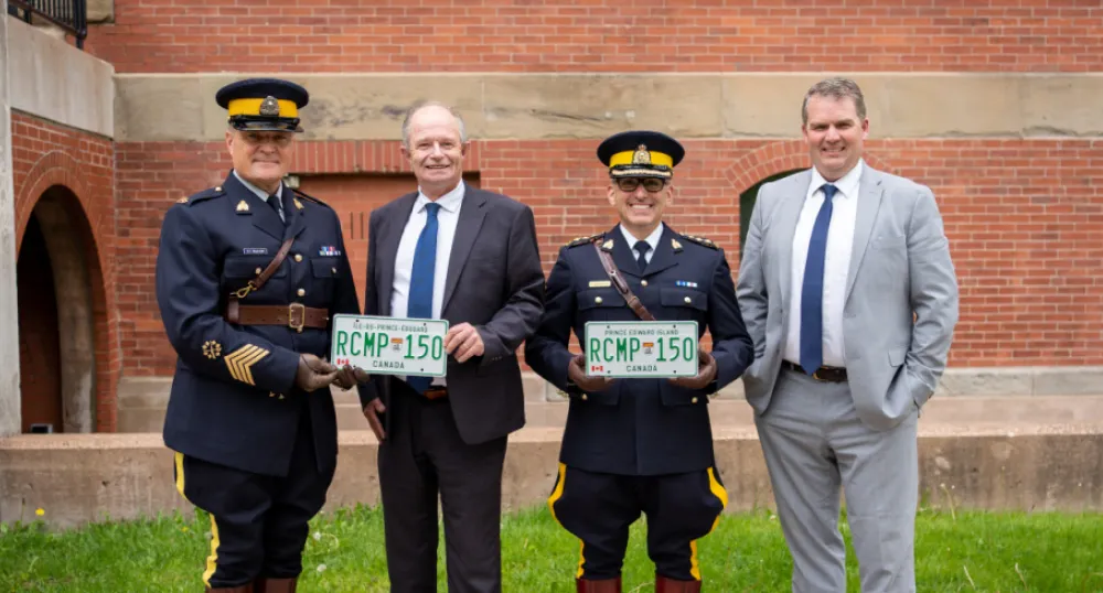 Province gifting RCMP with commemorative plates image of four people standing shoulder to shoulder in front of a building and holding a license plate