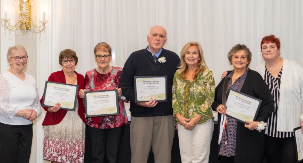 Province announces recipients of Senior Islander of the Year Awards for 2023 image of a group of people standing shoulder to shoulder in a room with a white curtain behind them and holding certificates in their hands
