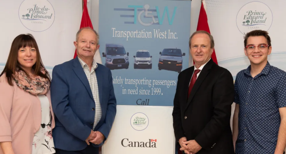 Residents of West Prince to benefit from a new fuel efficient and accessible vehicle fleet image pf four people standing shoulder to shoulder in front of a poster with vehicles on it.