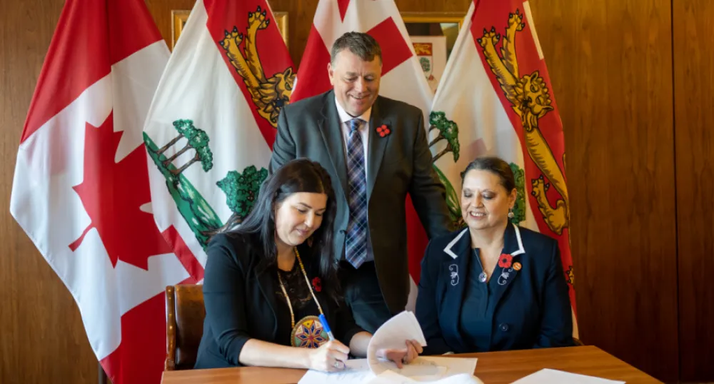 Prince Edward Island and First Nations sign Treaty Education MOU image of two people aitting at a table signing an agreement with another person standing behind them watching with some flags in the background