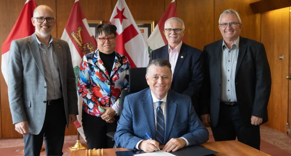 image of four people standing behind and overlooking a person while someone sits at the desk signing a proclamation