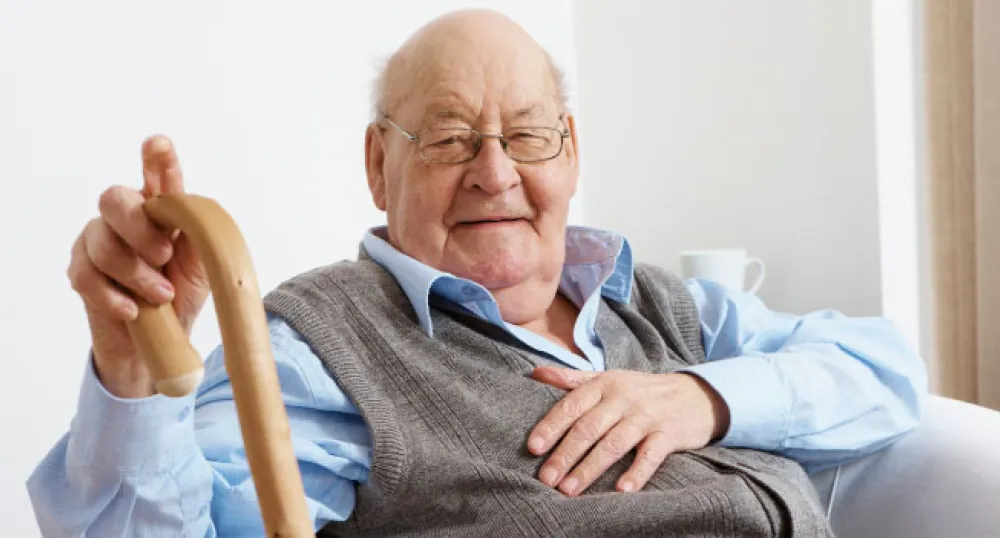 An elderly man with a cheerful expression sits comfortably in a chair, holding a cane in his right hand,