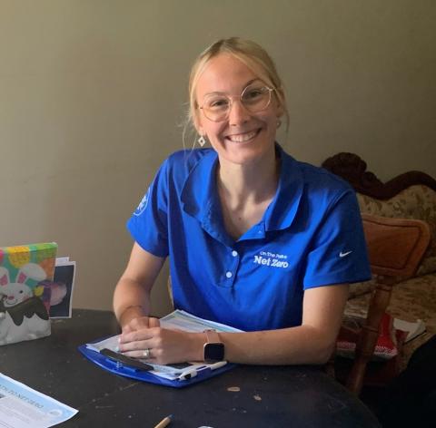 Julia Doiron, wearing a blue Net Zero t-shirt, sits at a desk with pamphlets in view on the desktop
