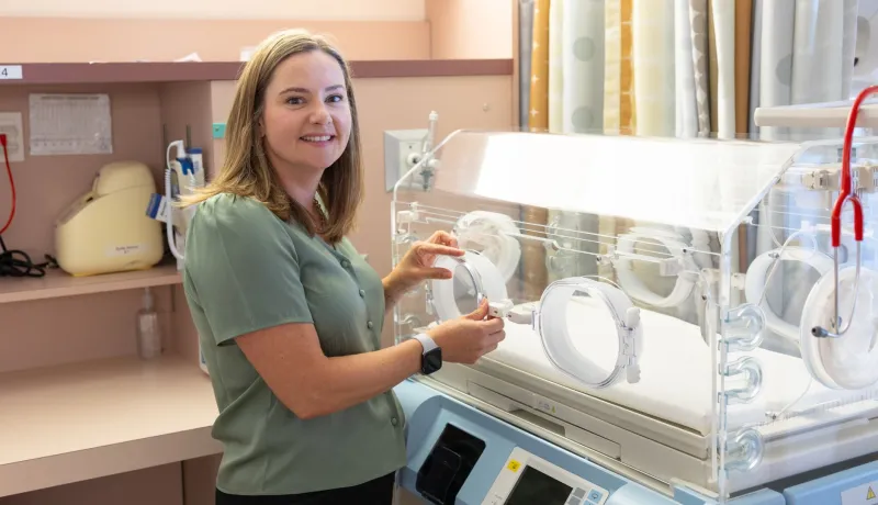 Dr. Maclean with shoulder length hair stands next to a NICU incubator.