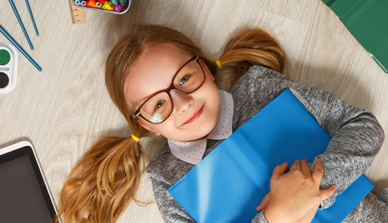 Young blonde girl with glasses laying on her back on the floor holding a book surrounded by books, pencils, paint and paint brushes