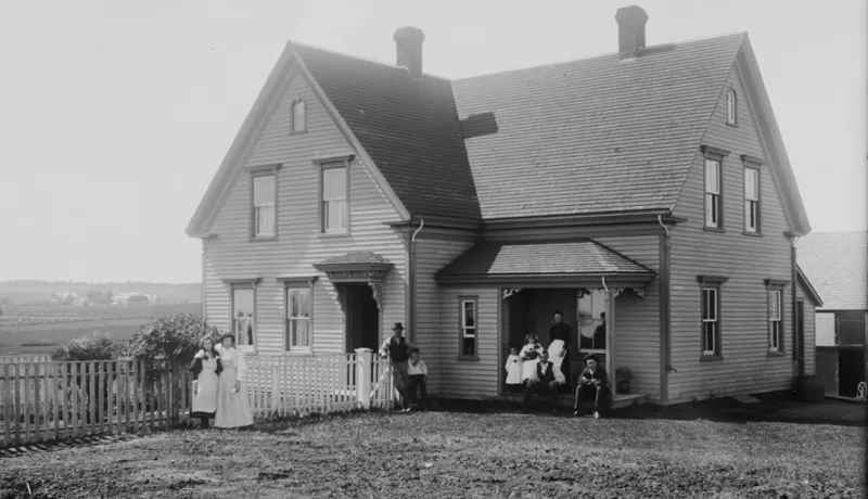 Exterior of the Brehaut house in Alexandra, Prince Edward Island, ca. 1890-1906. The family is posed on the front porch and in front of the fence. Rural landscape in background.