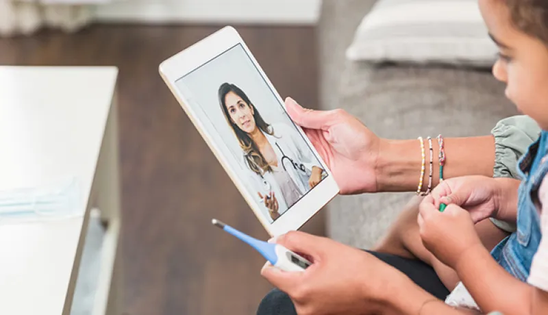 Child sitting on a mothers lap holding a thermometer and tablet speaking with a virtual care physician
