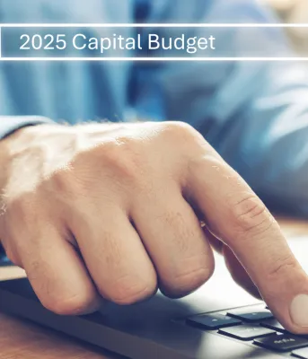 A person&#039;s hand resting on a laptop keyboard, with the screen displaying a search bar reading &quot;2025 Capital Budget&quot;