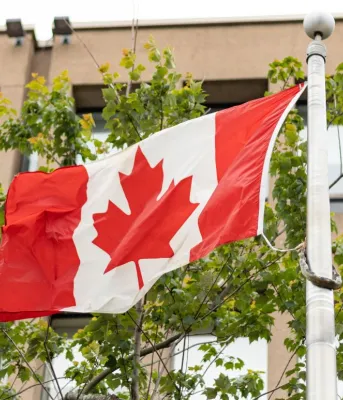 A Canadian flag blows in the wind with trees in the background.