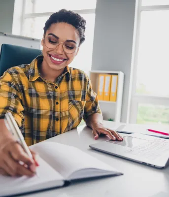 A person sitting at a desk holding a pen to a notebook and their laptop is open.
