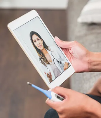 Child sitting on a mothers lap holding a thermometer and tablet speaking with a virtual care physician