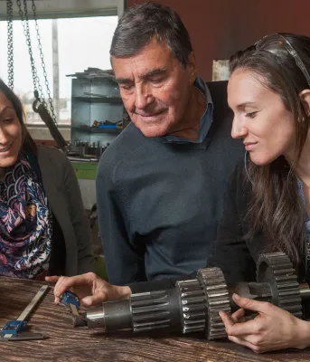 Young apprentice mentored by company founders Three people looking at a machine part and measuring its size.