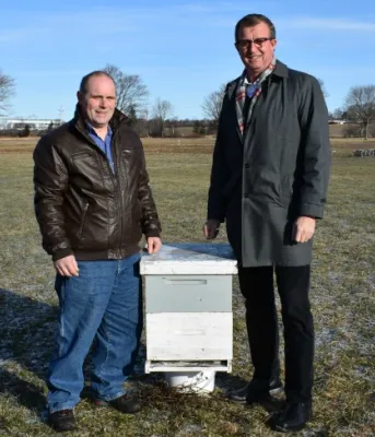 Province to increase pollination Photo shows two men standing on either side of a box-shaped beehive.