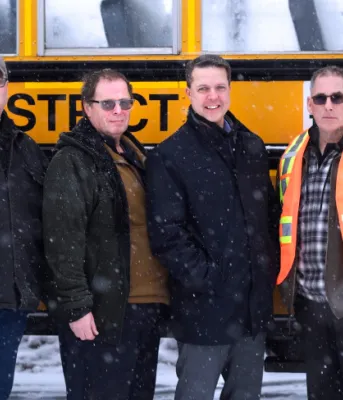 Minister Trivers and PSB Director Parker Grimmer stand outside beside a bus with school bus driver training participants