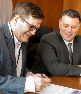 Two people sitting shoulder to shoulder at a desk signing a document.
