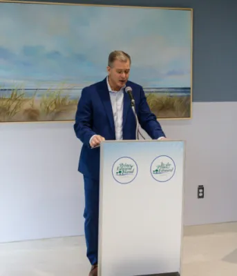 Minister McLane stands at the podium, three people seated to the right look on and one woman on the left is watching. Banners behind him show Island scenery.