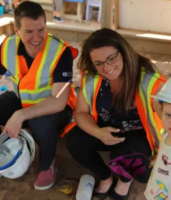 Minister Jordan Brown sits on the edge of a sandbox holding a hardhat, beside him sits Paige MacLaren, Director of the Morell Early Childhood Development Centre , and a cute little girl stands nearby.
