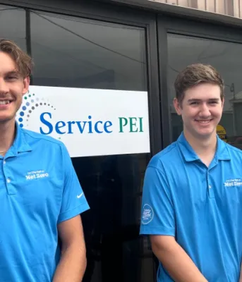 Three young men, Aaron Walsh, Johnny MacKenzie, Brandon MacQuillan, wearing blue Net Zero t-shirts stand in front of a Service PEI sign on a brick building. 