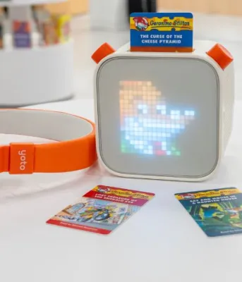 Families can borrow Yoto Audio Players at PEI public libraries image of orange coloured headphones and small cube audio player on a small round table